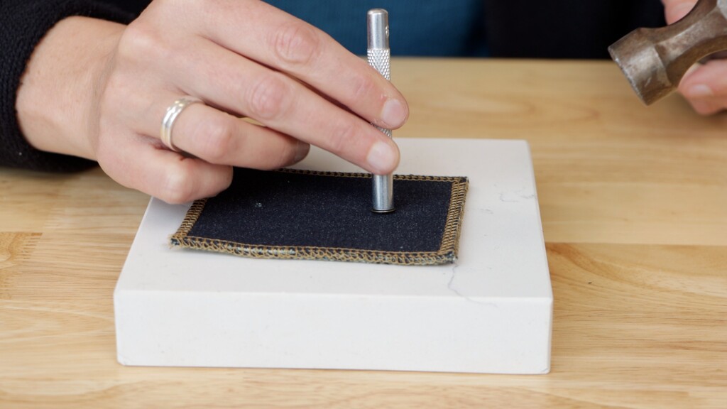 Hands using a hammer and setting tool to install a jeans rivet on a scrap of denim